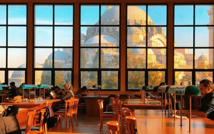 Interni accoglienti di una biblioteca con grandi finestre che mostrano una grandiosa architettura, e persone che leggono e studiano ai tavoli di legno.
