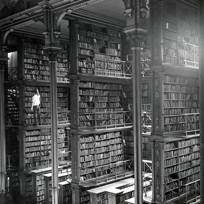 Interno grandioso di una biblioteca con imponenti scaffali, molteplici balconi e una persona che cerca un libro in un'ambientazione magica.