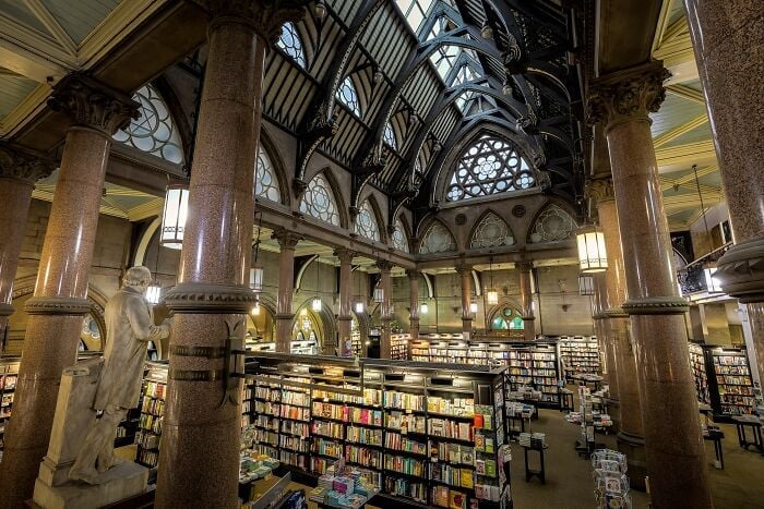 Interno grandioso di biblioteca con colonne imponenti, soffitti a volta e scaffali pieni di libri in un'ambientazione magica.