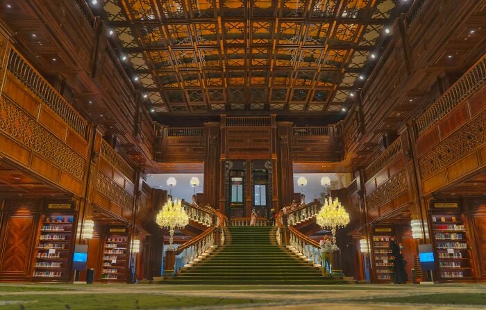 Interno grandioso di una biblioteca con una grande scalinata, pareti ornate in legno e scaffali pieni di libri in un'ambientazione magica.