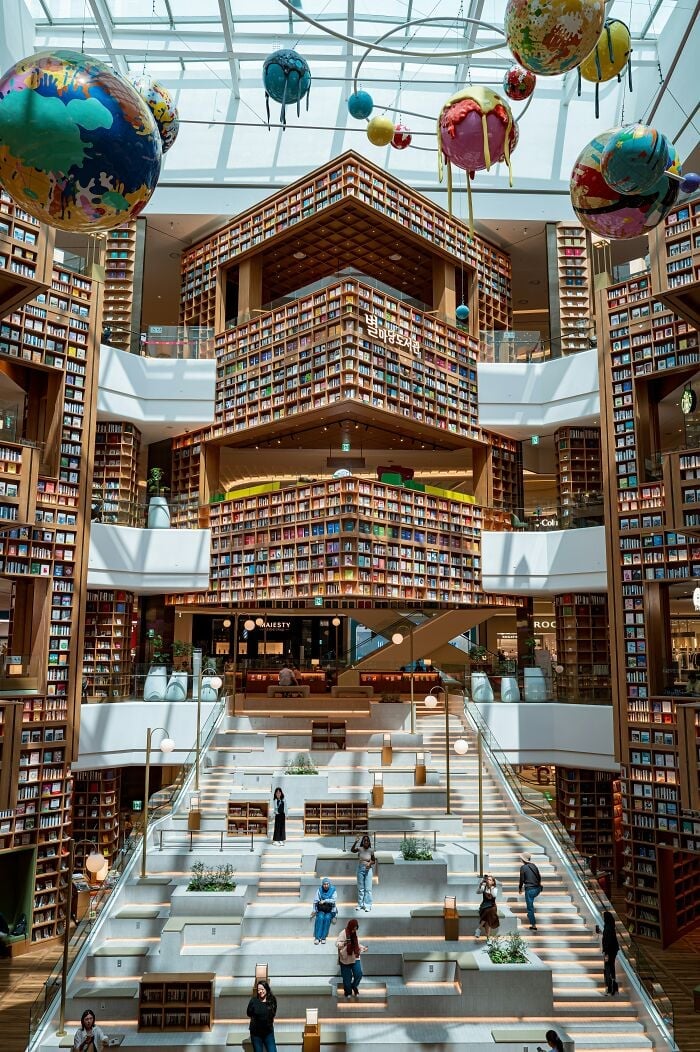 Interni grandiosi della biblioteca con imponenti librerie, grande lucernario e sculture di pianeti colorati appese al soffitto.