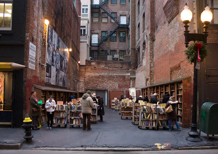 Outdoor cosy bookstore in an urban alley with people browsing books surrounded by brick walls and vintage street lamps.
